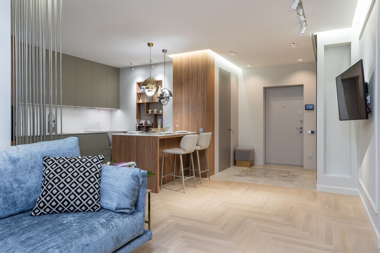 Interior of modern light studio with open kitchen with cabinets next to counter near chairs under lamps next to sofa with pillow in front of TV